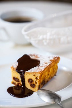 Kuru üzümlü, çikolata soslu, toz şeker ve naneli süzme peynir güveci. Beyaz bir antika tabakta duruyor, eski bir kaşık tabakta yatıyor. Tabak beyaz bir masa örtüsünün üzerinde. Masa örtüsünün üzerinde beyaz bir fincan kahve var. Büyük bir çömlek...