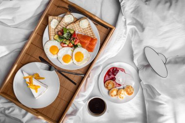 Fried toast with salmon slices, avocado, cucumbers, sesame seeds, tomatoes, fried egg and butter with parsley, cheesecake with apricot slices on a wooden board cheese pies in a plate with sour cream and jam on a white table