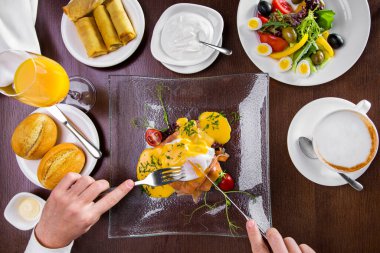Breakfast at the hotel from egg benedict with salmon, cheese sauce, cherry tomatoes, microgreen in a glass, transparent plate. Male hands are holding a fork with a knife. To the right of the plate is a cup of cappuccino. Above is a white plate of qua
