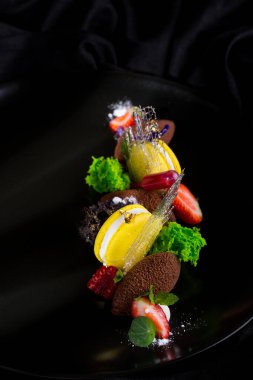 Macaroni with pastries, strawberries, sugar thread and salad on a black plate and a dark background. Vertical orientation