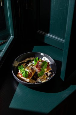 Sandwiches with cheese, meat, basil, almonds, kumquat, sauce and sesame seeds in a black plate against a dark green window background