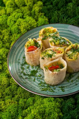 Domatesli lavaş, salatalık, kabak, kırmızı soğan, patlıcan ve maydanoz, ızgara ve rulo. Parça parça dilimlenmiş. Lavaş yuvarlak, mavi, seramik bir tabakta yatıyor. Plaka yeşil ve açık yeşil yosun arkaplanının karşısında duruyor..