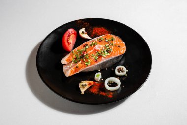 Raw salmon steak with peppercorns, tomatoes, onions, paprika, salt, thyme, and garlic in a plate on a white background