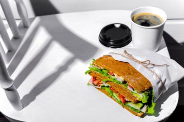 Sandwich in white paper with ham, lettuce, tomatoes, cucumbers and sauce with a paper cup of cappuccino and a black lid standing on a white chair against a background of a white wooden wall