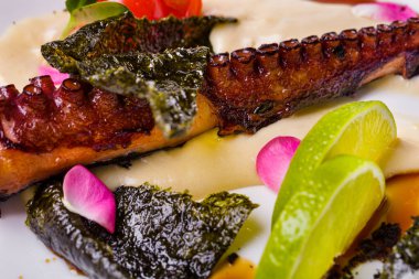 Mantar soslu kızarmış ahtapot, limon dilimleri, unagi sosu, vişneli domates ve mikroyeşil, beyaz tabakta. Çatallı beyaz masa örtüsü, ikinci bir tabak ve bir bardak gül şarabı..