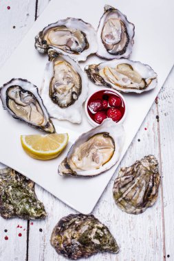 Okyanus istiridyeleri, üst kapakları olmadan, beyaz, dikdörtgen bir tabakta. Yakınlarda bir dilim limon ve çilek reçelli bir tencere var. Çanak çömlek hafif ahşap bir zemin üzerinde duruyor, deniz kabuklarının kapakları yakınlarda duruyor..