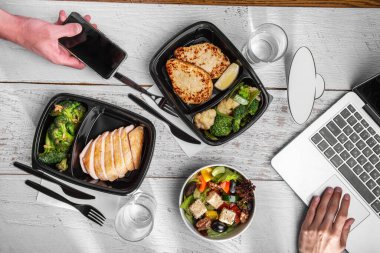 Kızarmış brokoli, limon, susam tohumu, feta peyniri, domates, zeytin, kızarmış pirzola, marul, ceviz ve biberli çatal bıçak, dizüstü bilgisayar, su bardakları ve beyaz ahşap masa üzerinde telefonu olan erkek eli.