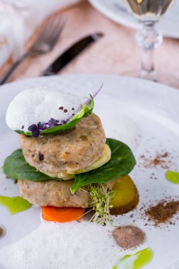 Steamed fish cakes with spinach leaves, knee cucumber, carrots and citrus foam. The fish is on a white ceramic plate. The plate is on a stand with a beige tablecloth, next to it is a cloth napkin, a fork with a knife, two glasses of white wine and an