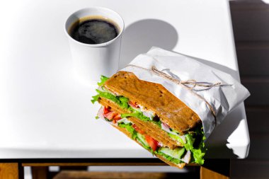 Sandwich in white paper with ham, lettuce, tomatoes, cucumbers and sauce with a paper cup of espresso standing on a white chair against the background of a white wooden wall