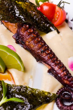 Mantar soslu kızarmış ahtapot, limon dilimleri, unagi sosu, vişneli domates ve mikroyeşil, beyaz tabakta. Çatallı beyaz masa örtüsü, ikinci bir tabak ve bir bardak gül şarabı..