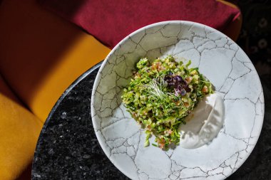 Oriental salad appetizer Tabbouleh with quinoa, finely chopped parsley, cucumber, tomato and microgreens. Tabbouleh lies in a light round ceramic plate with high sides. The plate sits on a black table covered in sparkles. Nearby is a yellow armchair 