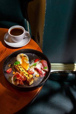 A slice of cheesecake with thyme, prosciutto, parmesan, basil, tomatoes, garlic and a boiled egg in a plate on the table with a cup of coffee