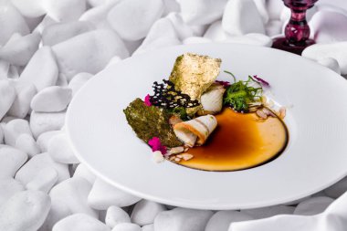 Tütsülenmiş yılan balığı ve balzamik soslu panna cotta, üstünde chuka salatası, badem gevreği, yenilebilir taç yaprakları, yaldızlı Norveç yosunu. Yemek derin beyaz bir tabakta. Tabak beyaz taşların arka planında duruyor..