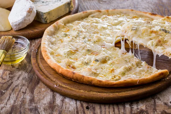 Pizza with different types of cheese on a wooden table being lifted with a spatula and lying on the table with four types of cheese lying aside on a wooden board