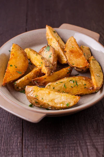 Bol yağda kızartılmış patates dilimleri, sert tuzlu, üzerine doğranmış maydanoz serpiştirilmiş. Patatesler seramik tabakta. Tabak koyu ahşap bir arka planda duruyor..