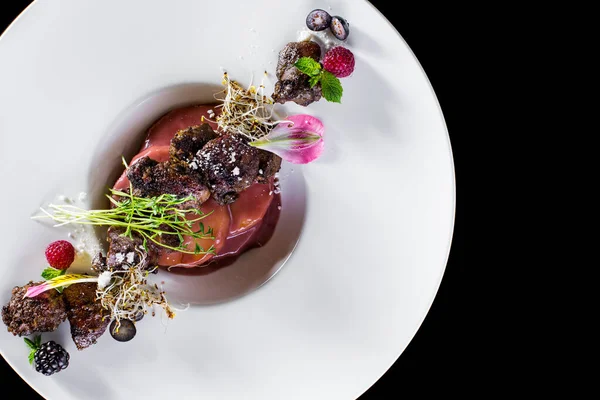 Grilled foie gras with pineapple in cherry sauce, microgreen sprouts of wheat and onion, blackberries, raspberries and blueberries, edible petals. Foie nra lies in a round ceramic plate. The plate stands on a black background.