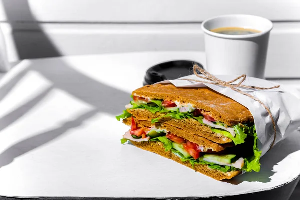 Sandwich in white paper with ham, lettuce, tomatoes, cucumbers and sauce with a paper cup of cappuccino and a black lid standing on a white chair against a background of a white wooden wall