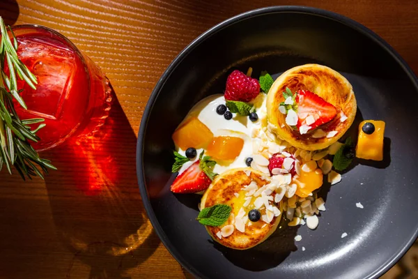 Cheese pancakes with mango, strawberry, raspberry, almond slices, mint and sauce in a plate on the table with a glass of juice and rosemary