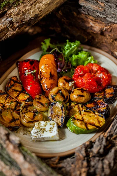 Grilled vegetables on the grill, red paprika, yellow paprika, zucchini, mushrooms, onion, tomato, with lettuce leaves, olive oil, feta cheese and herbs. They lie on a ceramic, oval plate, among the wooden logs.