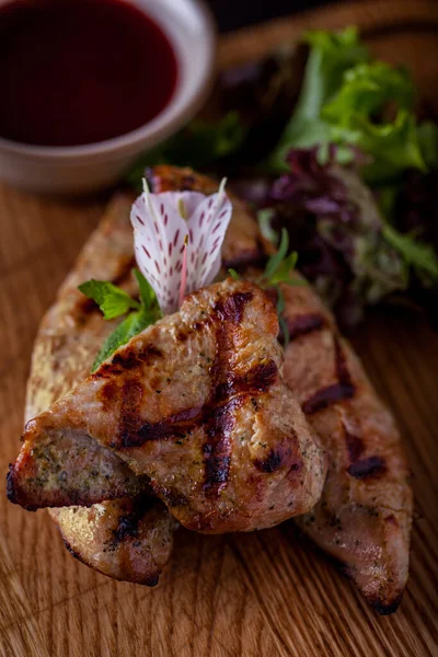 Grilled chicken fillet shashlik. Nearby is a beige sauce bowl with cranberry sauce, a salad of several types of lettuce. Food and sauce stand on a wooden board.