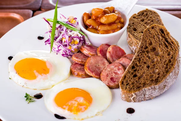 Scrambled eggs from two eggs and grilled sausages cut into circles. Purple cabbage and corn salad with green onion feathers. On the plate there is a gravy boat with beans in a tomato. Balsamic drops on a plate. The plate stands on a wooden background