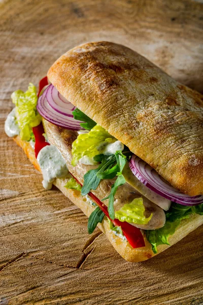 Tavuk sosisli Chiabatta sosisi, marul, taze soğan halkaları, taze paprika, sarımsak sosu, roka. Ahşap bir arka planda yatıyor.