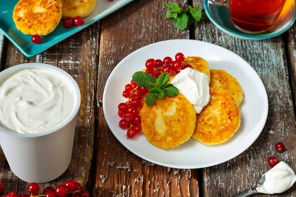 Cheesecakes with mint, sour cream and red currants in a plate on a wooden table with a spoon and sour cream in a glass