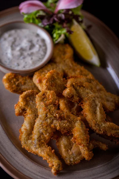 Fried frog legs in batter with sauce, lemon, salad and flower petals on an oval plate on a wooden table. Vertical orientation