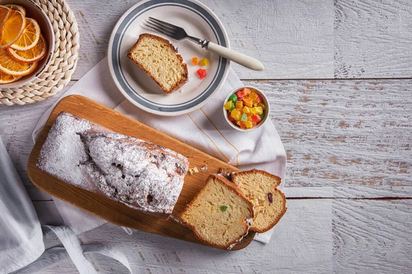 White sweet bread with powdered sugar cut into pieces with dried oranges lying on a wooden board with a towel and a fork on the table