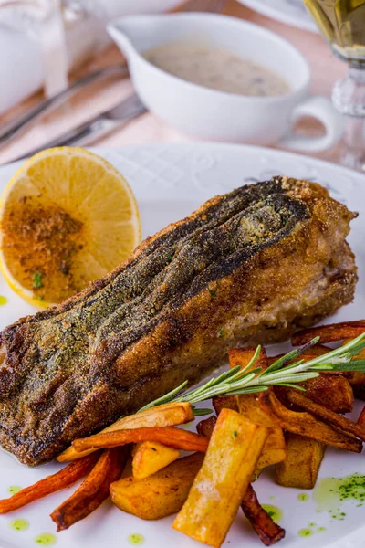 Fried fish steak, breaded with lemon slices, rosemary sprig, fried fries, white sauce, on a white plate. Vertical orientation 