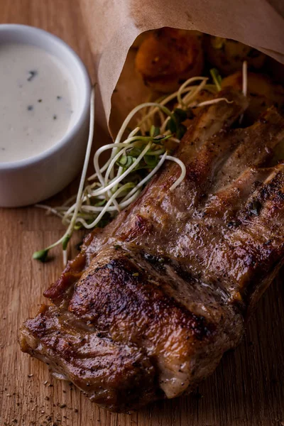 Grilled pork ribs with baked potatoes and microgreen pea sprouts, lie in a paper envelope. Next to it is a white gravy boat with garlic sauce. The food and sauce are on a wooden board, next to a large glass of light beer.