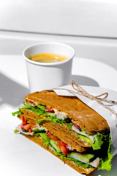Sandwich in white paper with ham, lettuce, tomatoes, cucumbers and sauce with cappuccino with a paper cup standing on a white chair against the background of a white wooden wall