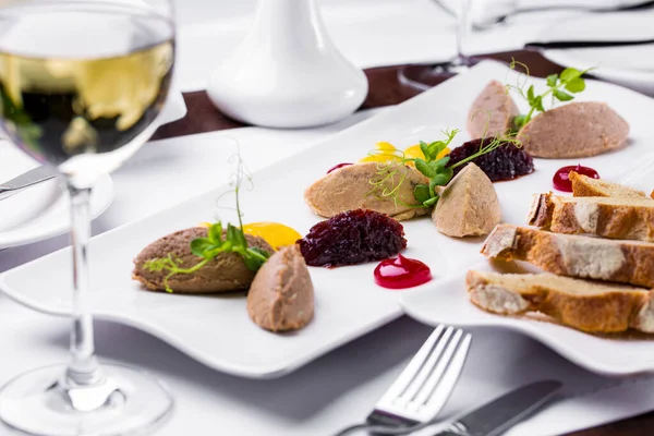 Assorted rabbit liver and chicken liver pate, glazed onions, pea microgreen sprouts and orange and cherry marmalade next to it. The pate lies on a white, rectangular plate. Nearby is a plate of toast, a glass of white wine, a fork and a knife, a vase