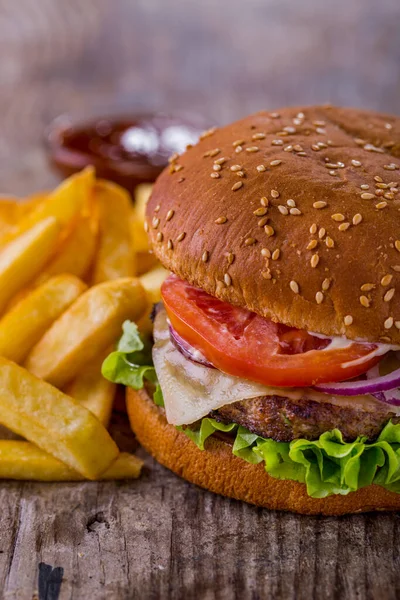 Burger with sesame bun, cutlet, tomatoes, onions, lettuce, cheese and mayonnaise with fries and sauce on a wooden table. Vertical orientation