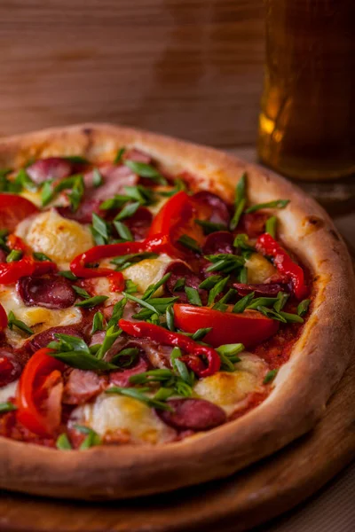 Pizza with cherry tomatoes, ham, sausages, green onions and cheese on a wooden board with a glass of beer on the table