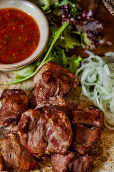 Grilled pork skewers, lying on pita bread, next to lettuce and arugula salad and sliced onion rings. A sauce-pot with adjika lies nearby, Food and sauce are on a wooden board. The board stands on a wooden background with acacia logs. A glass of red w