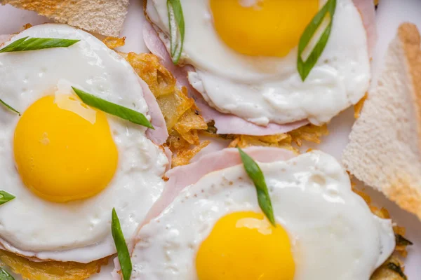 Breakfast of three fried eggs, fried eggs, on top of the ham. The ham and eggs are on top of the potato grills. Nearby are white bread toasts. Sprinkled with chopped green onions on top of the eggs. The food is on a white plate. The plate is on a lig