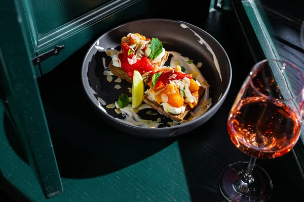 Sandwiches with indal, lemon, mint, apricot, pickled pepper and white sauce in a black plate against the background of a dark green window with a glass of wine