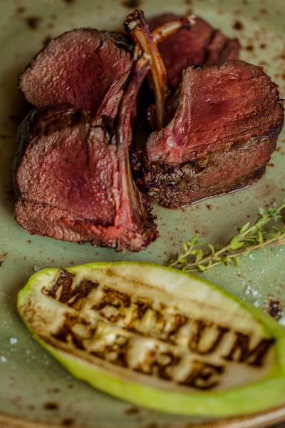 Rack of veal, medium rare, sliced in portions. Next to it is a sprig of thyme and a slice of zucchini with a roast signet. The food is on a green ceramic plate. The plate is on a wooden board, next to it are metal dishes for oil and spices, a glass o