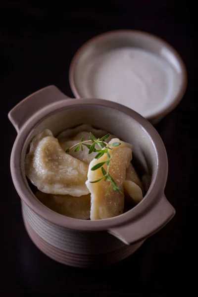 Homemade, boiled, dumplings with potatoes and oven baked in a ceramic pot with a rosemary branch. Nearby is a small bowl of sour cream. Black background.