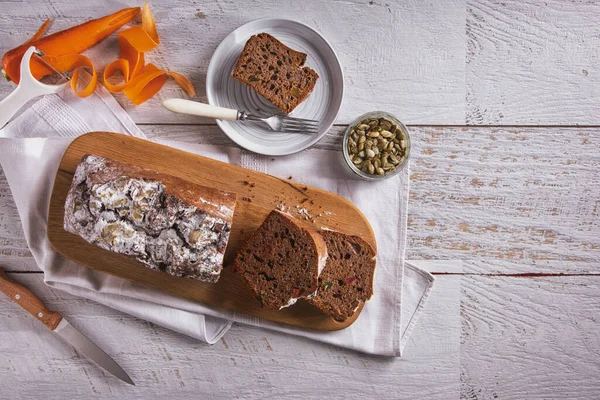 Sweet bread with powdered sugar, carrots and watermelon seeds, cut into pieces, lying on a wooden board with a towel and a fork on the table