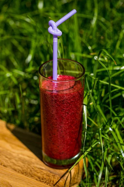 Raspberry smoothie in a tall and transparent glass on a wooden board among the lawn in the sunlight.