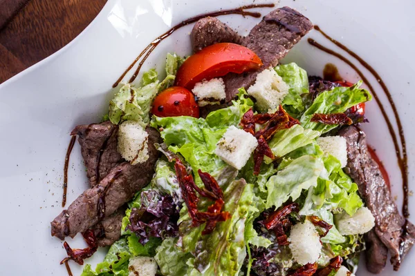 Salad with beef meat, lettuce, sauce, sesame seeds, endive, croutons, tomatoes and arugula in a plate on a wooden background