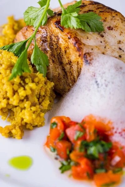 Maydanoz, biber, süt köpüğü, kuskus ve soslu kızarmış dorado bifteği hafif arka planda beyaz yuvarlak bir tabakta. Dikey yönelim