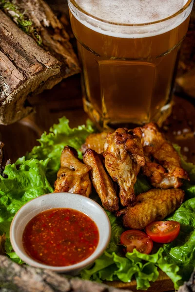 Grilled chicken wings with cherry tomatoes and lettuce, next to it there is a gravy boat with tomato sauce. There is a glass of beer behind. Food and beer stand on a wooden board. There are barbecue logs around.
