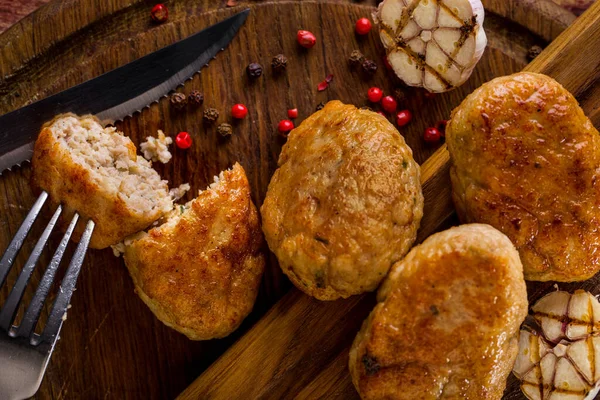 Four chicken cutlets lie on a wooden board. One cutlet is cut in half, a fork is stuck in half. There is a knife on the board, next to them are peppercorns and grilled garlic.