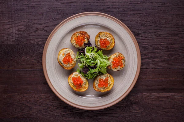 Patatesler dilimlenip fırında kremalı sos ve dereotla pişirildi. Her patatesin üstünde kırmızı havyar var. Patatesler seramik üzerinde, yuvarlak tabakta, ortada roka ve marul salatası karışımı duruyor. Tabak ahşap bir arka planda duruyor..