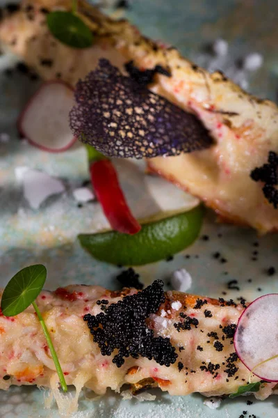 Kamchatka crab claws baked with cheese. On top are black caviar, microgreen sprouts and edible violet flowers. Next to it is a mango sauce, radish slices and lime wedges. The food lies on a ceramic, circular plate with a blue pattern. The plate is on