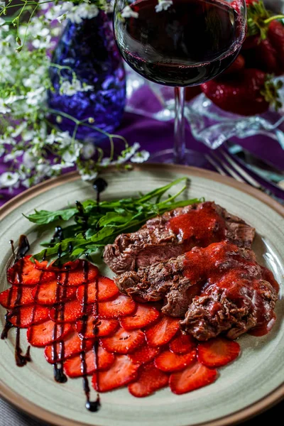 Fried pieces of meat with strawberries, strawberry jam, herbs, flowers on a round plate and with a glass of tap wine. Vertical orientation