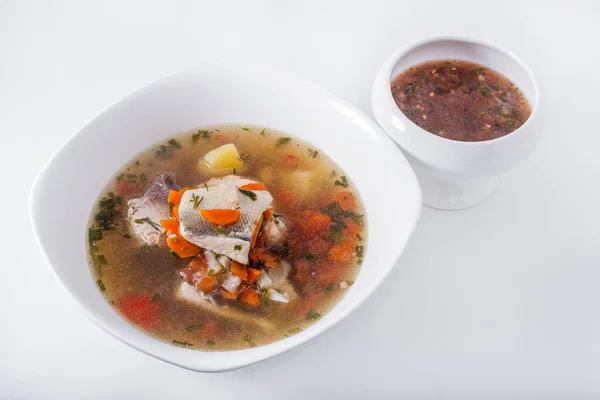 Soup with fish, potatoes, dill, parsley, carrots and peppers on a plate with sauce and a white background